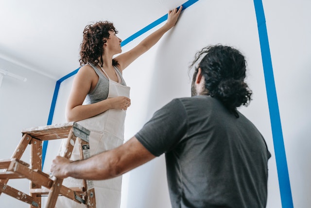 A-person-standing-on-a-ladder-painting-a-wall-while-another-person-holds-the-ladder-1